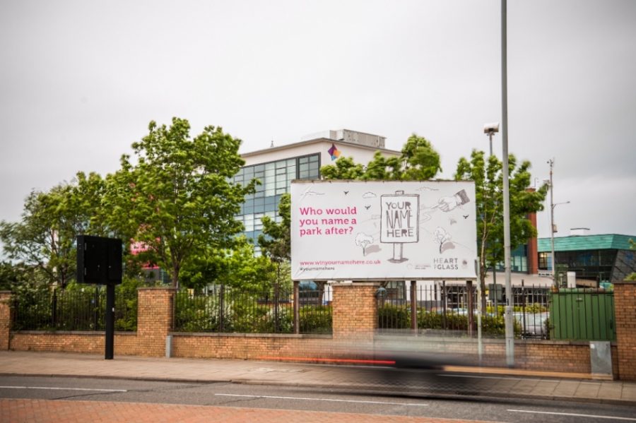 In daylight a large billboard stands at the side of a road. The billboard stands above a brick and metal fence. Behind it are some trees and the top of a large building with many windows. The billboard includes a grey background with pink text which reads 'Who would you name a park after?' as well as an illustrated sign which reads 'Your Name Here'