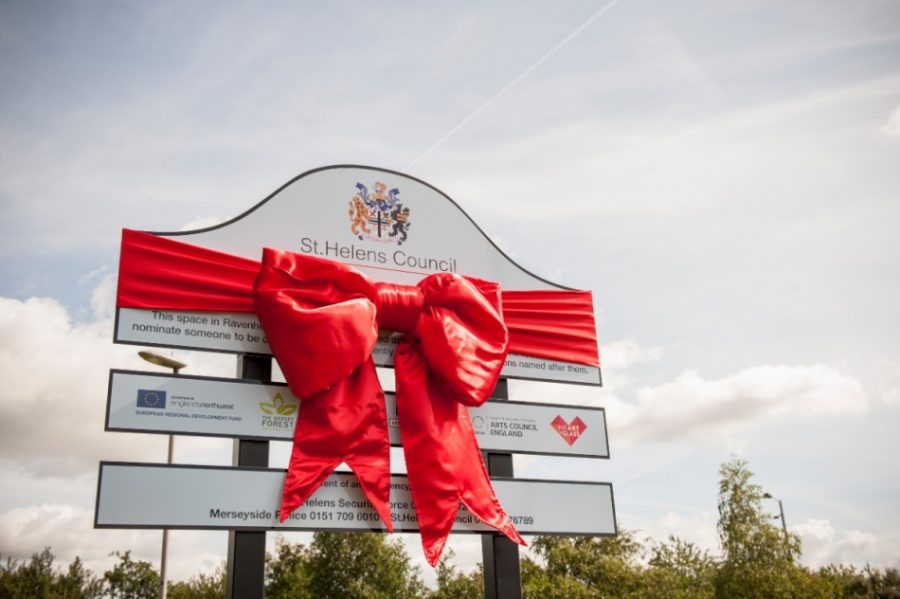 In daylight a large sign stands against a light blue sky with clouds and a line of tree tops underneath. The sign is partially covered by a large red, shiny bow. At the top of the sign you can see a crest and the words 'St Helens Council'.