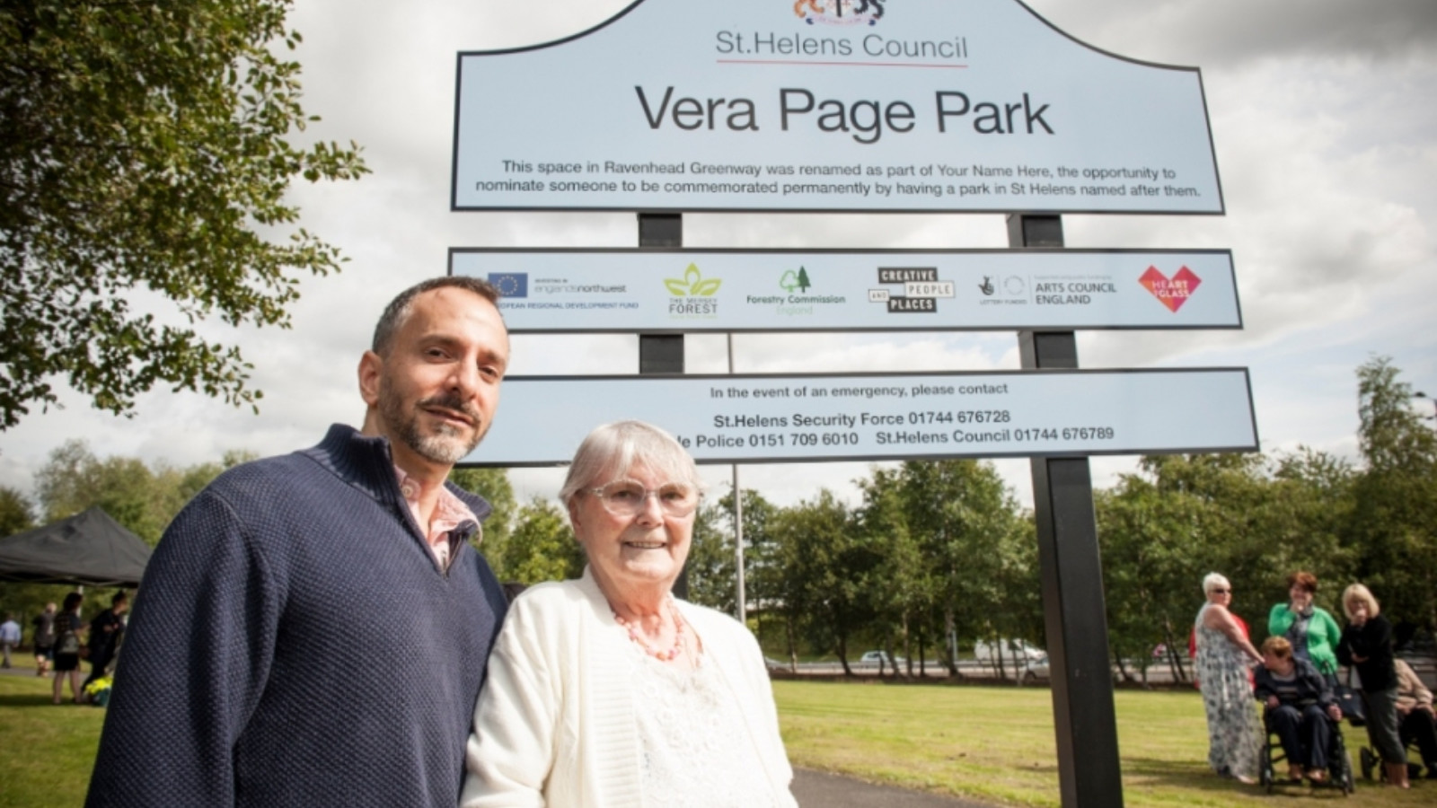 Two people, one older woman and one man, stand with their arms linked in front of a large sign. The sign has a crest at the top and reads 'St Helens Council. Vera Page Park' in large letters.
