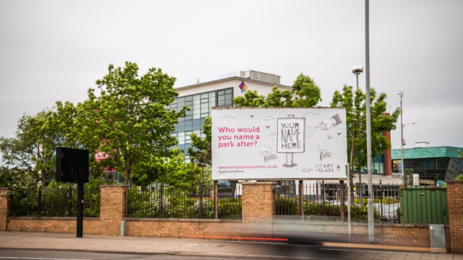 In daylight a large billboard stands at the side of a road. The billboard stands above a brick and metal fence. Behind it are some trees and the top of a large building with many windows. The billboard includes a grey background with pink text which reads 'Who would you name a park after?' as well as an illustrated sign which reads 'Your Name Here'