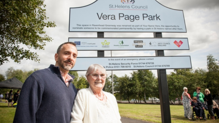Two people, one older woman and one man, stand with their arms linked in front of a large sign. The sign has a crest at the top and reads 'St Helens Council. Vera Page Park' in large letters.