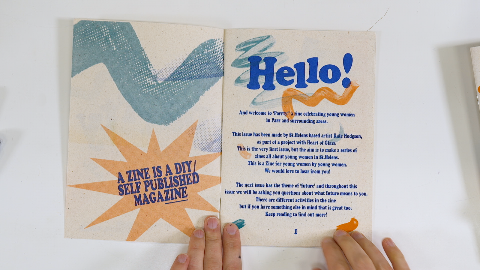 Two hands holding a zine open. The open spread displays printed text and abstract shapes. The word 'Hello' is written in large, bold, bright blue lettering