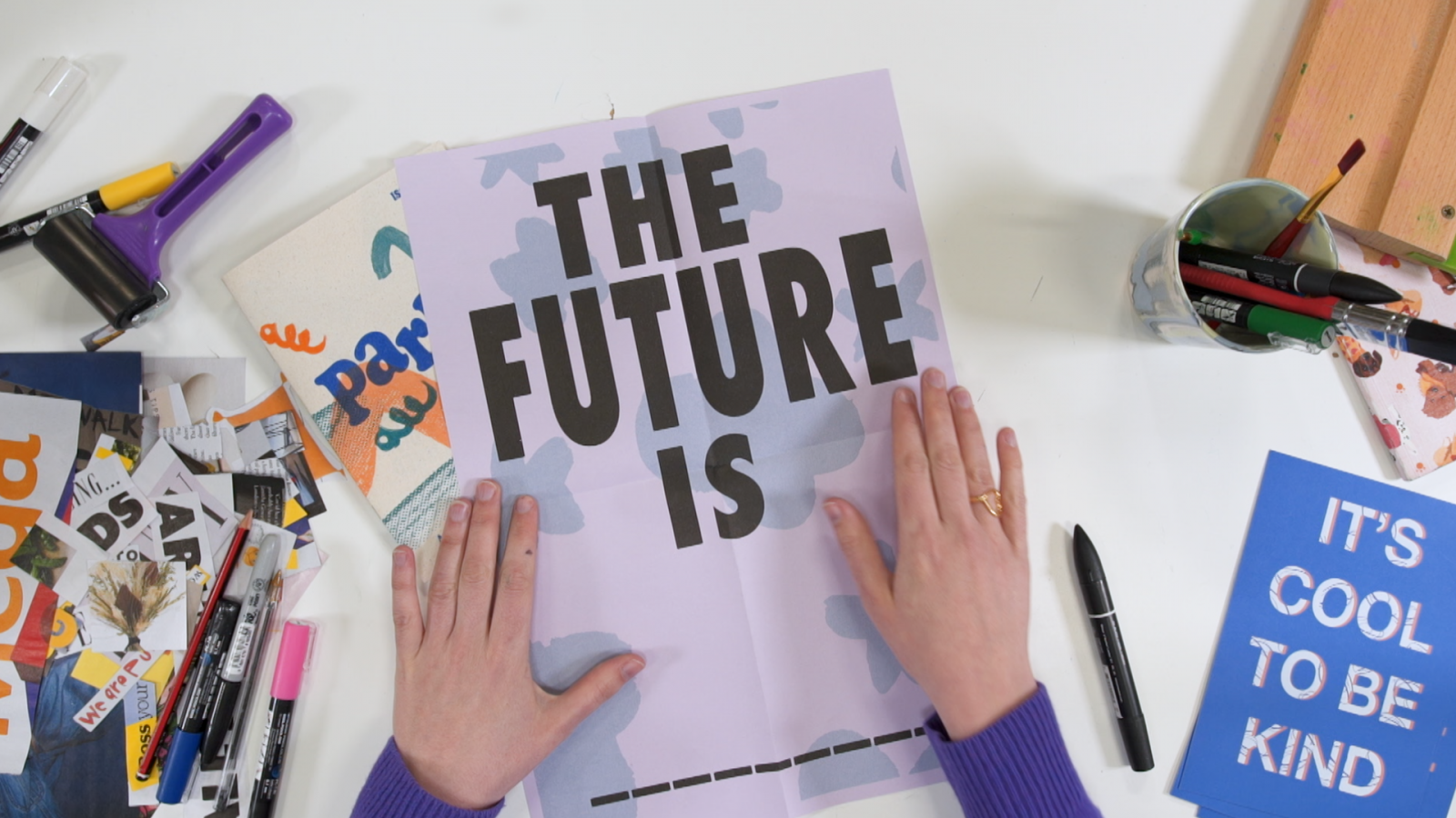 Two hands holding an A3 sized poster flat against a table. The poster is lilac, punctuated with light blue abstract shapes. The words 'The Future Is' are written in bold black capitals across the top of the poster.