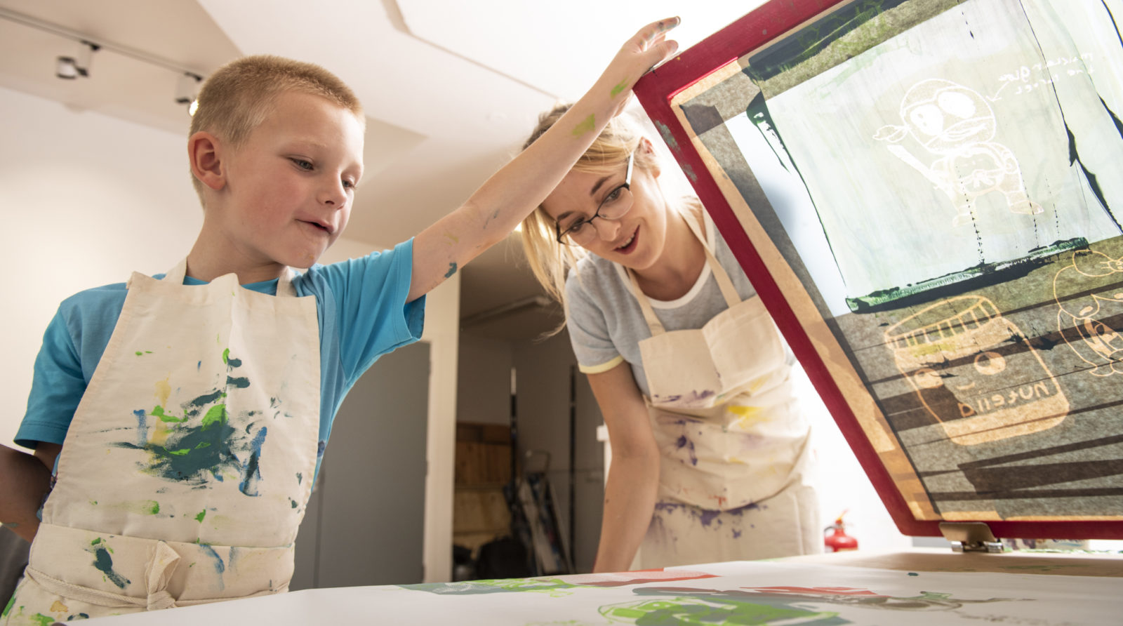 A young boy and a women are screen printing together. Both wear white aprons splattered with paint.