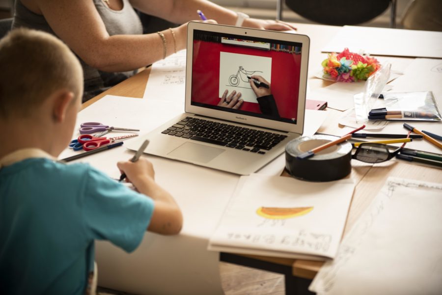 A table has lots of paper, pens and pencils on it and a laptop showing someone drawing a motorbike. A young person sits at the table and is drawing.