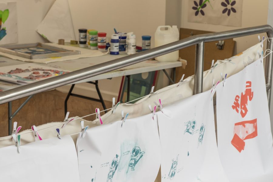 A white room has a table with arts supplies and paper on. The room is full of screen printed posters hanging on the walls and on string.