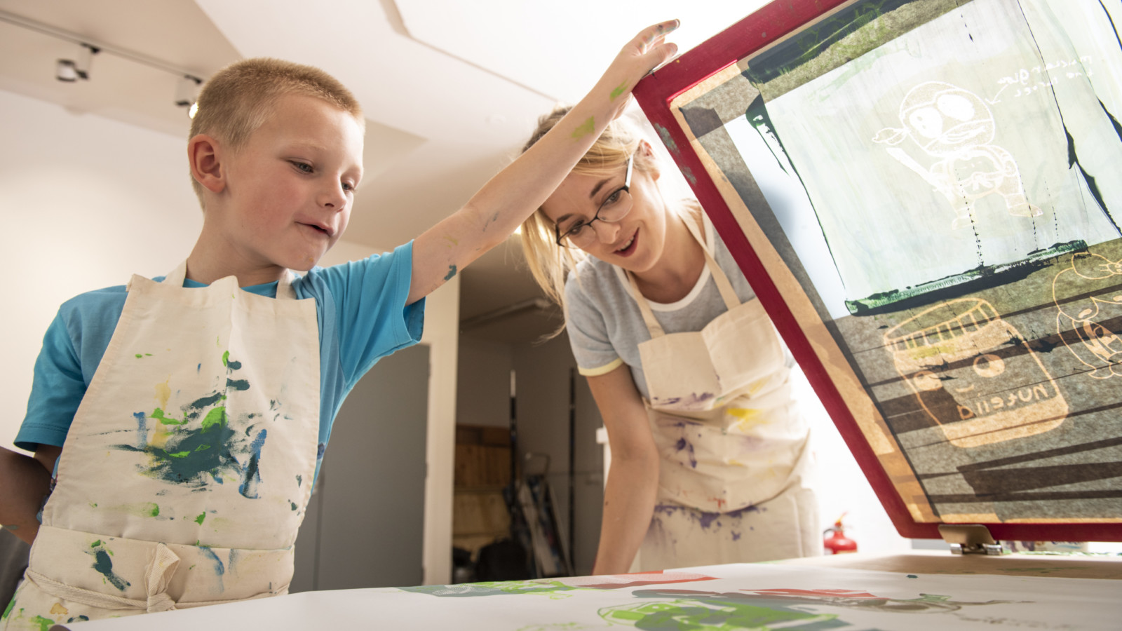 A young boy and a women are screen printing together. Both wear white aprons splattered with paint.