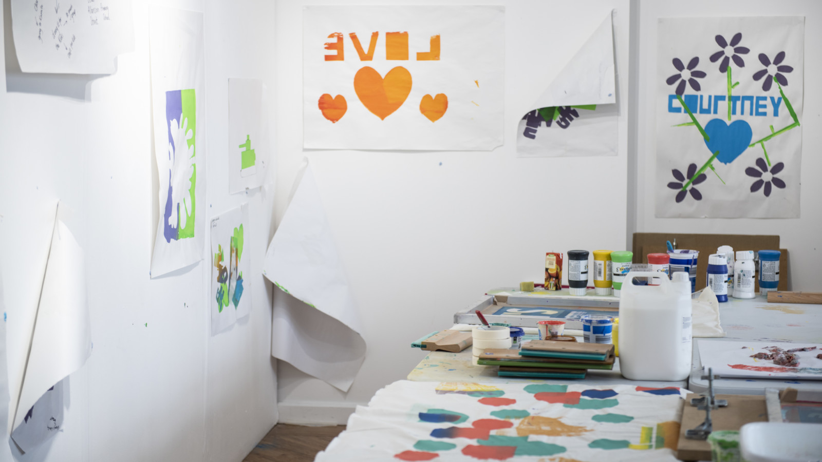 A white room has a table with lots of art supplies and paintings on. On the walls are posters with colourful paintings or hearts and flowers.