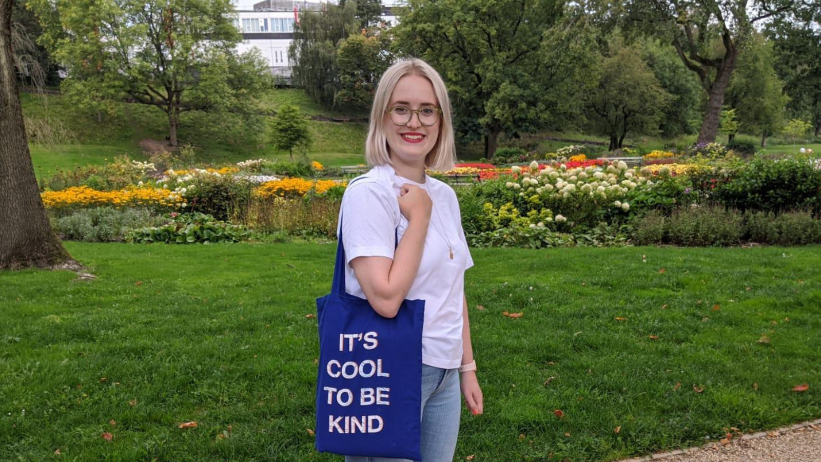 A woman in a white t-shirt and jeans stands in a large garden. A blue canvas bag with the words 'It's cool to be kind' printed in capital letters printed in white letters.
