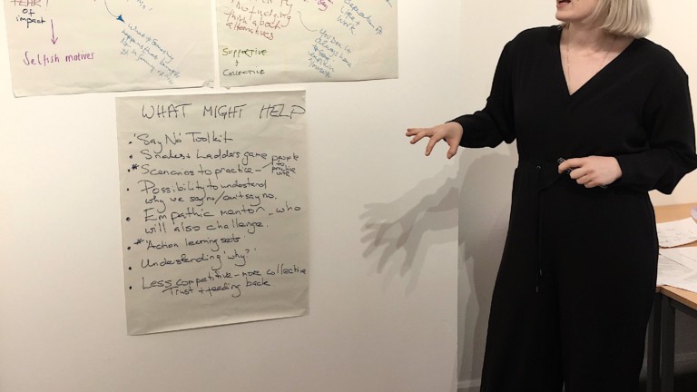 A woman with a light blonde bob and wearing a black jumpsuit stands looking at 3 mindmaps on the wall behind her.