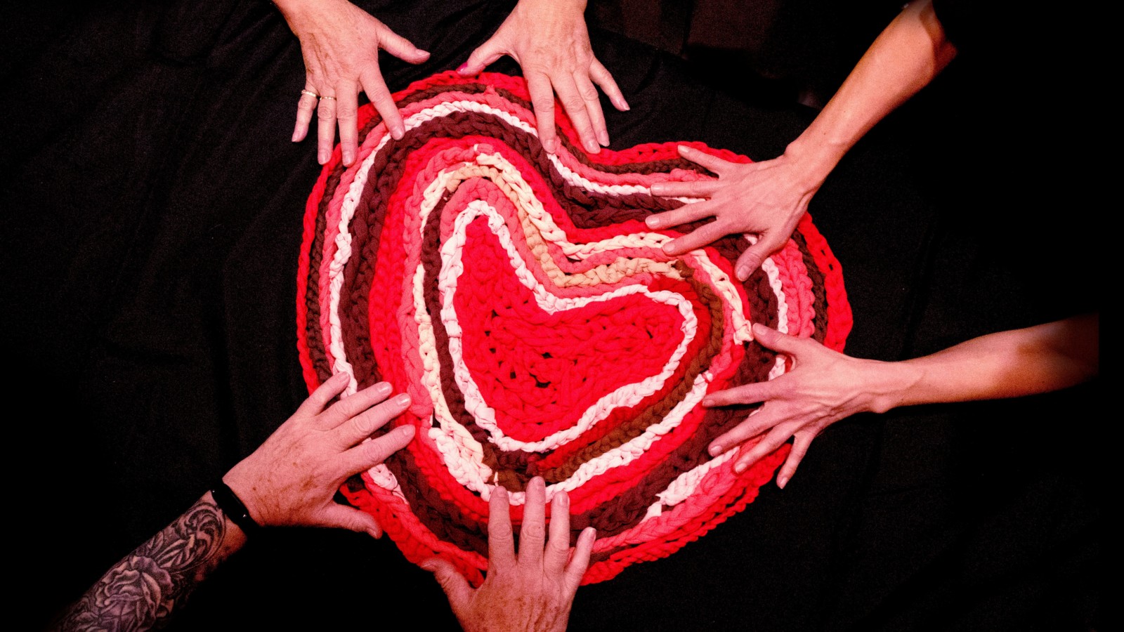 On a black background three pairs of hands are laid on top of a red and white knitted heart