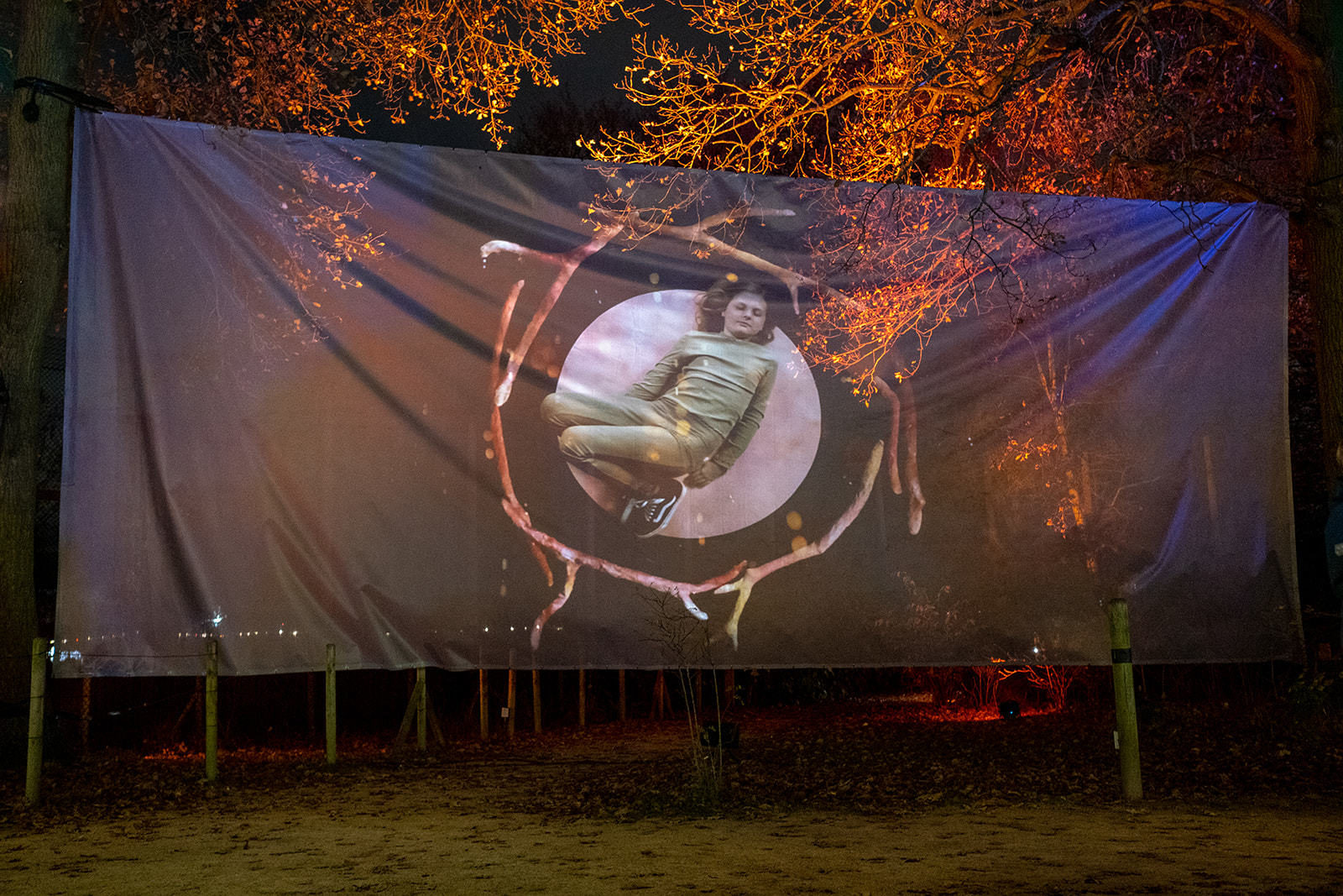 A large piece of fabric hangs between two trees. On it is an image of a girl projected. She is lying down with her eyes closed, a circle of antlers around her.