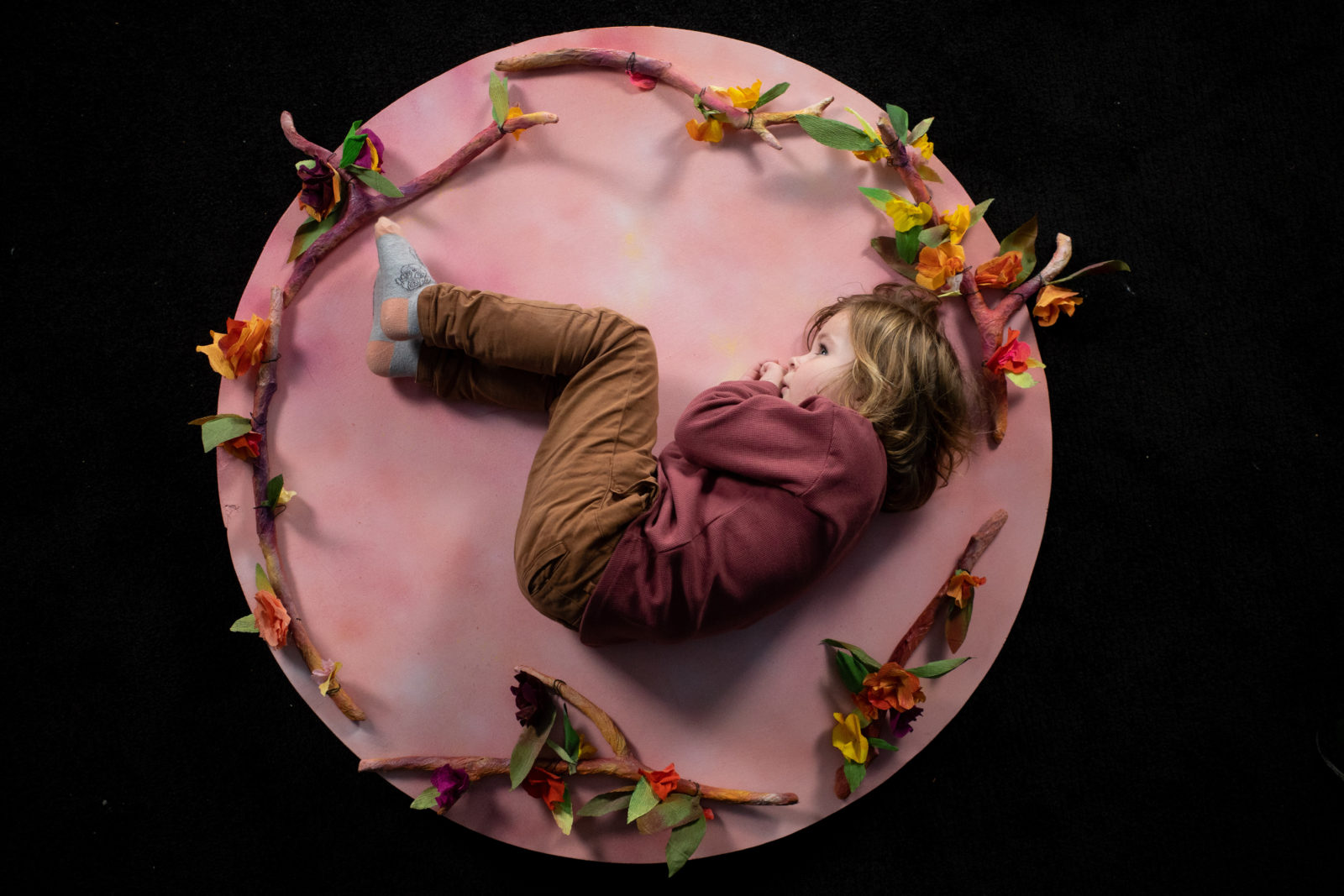 Child lying curled up on large pink hand made moon, surrounded by hand made floral antlers.