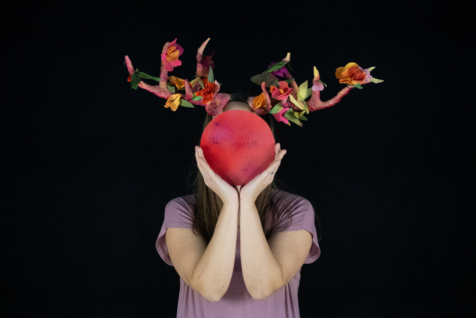 Woman wearing hand made floral antlers, holding hand made red moon covering her face.