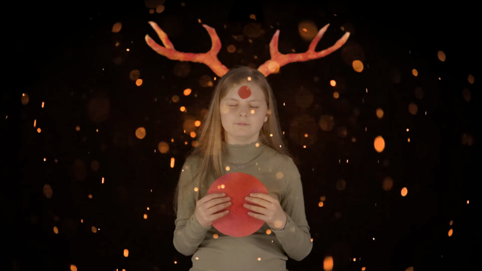 A girl wearing hand made antlers with a red circle on her head and in her hands, surrounded by orange glowing speckles.