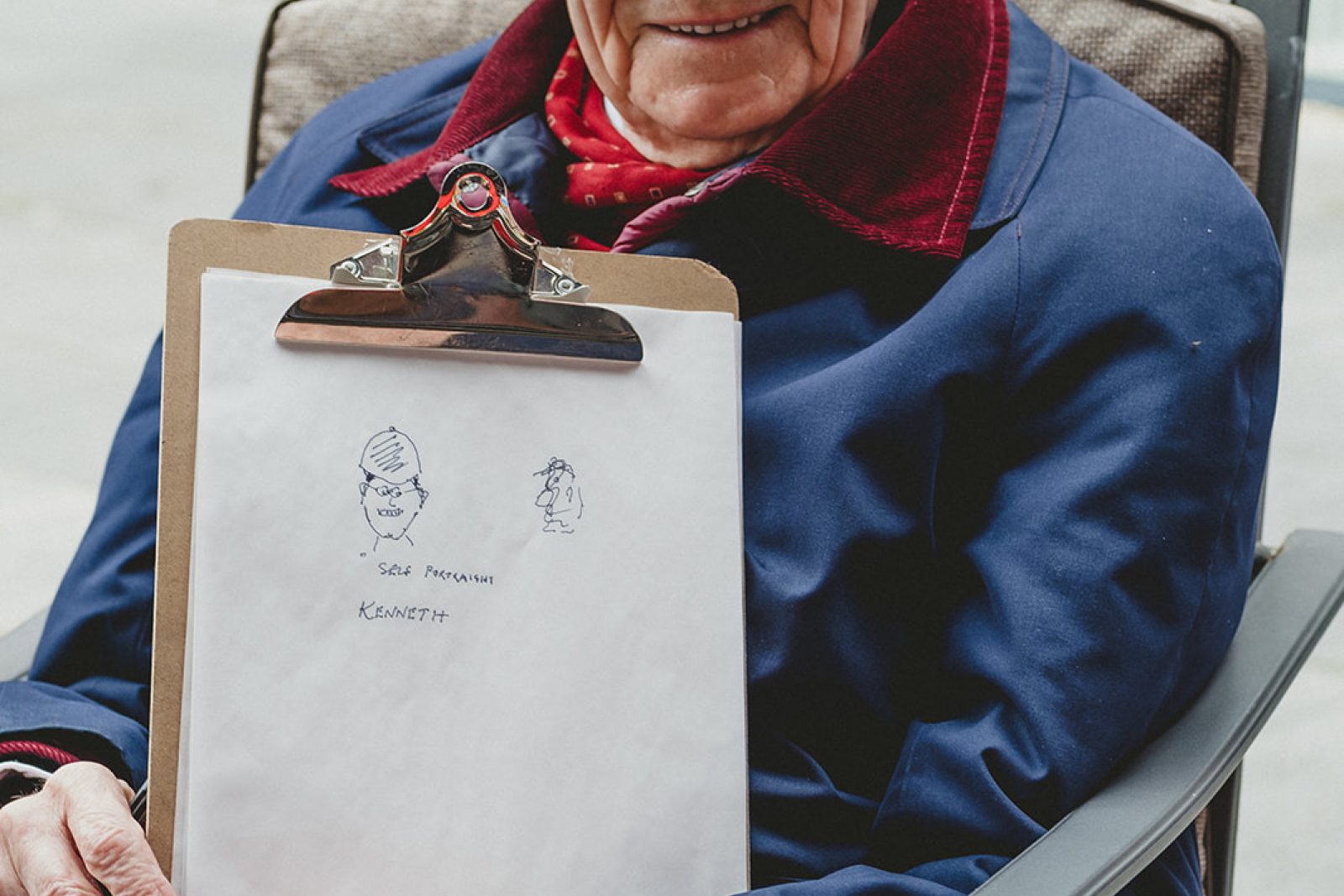 An older man wears tweed hat and a dark blue coat with a red collar. He holds a clip board containing paper with a drawing of two faces.