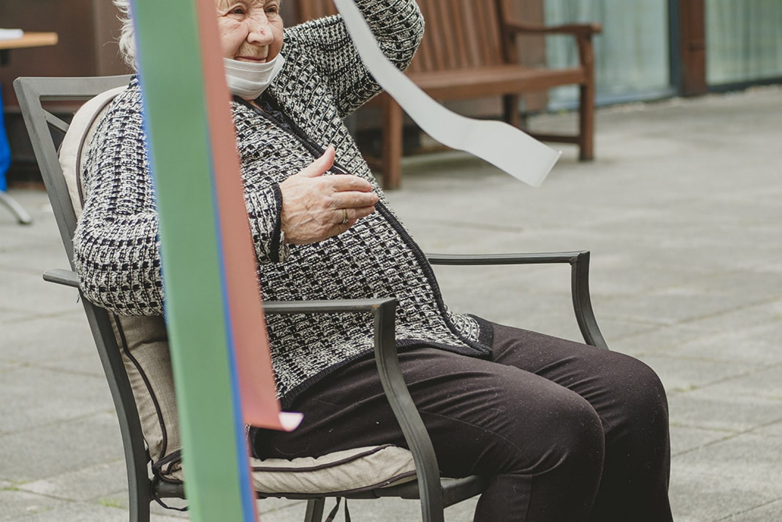 An elderly woman is sat in a chair. Her left arm is raised above her head and her right arm is held in front of her body.