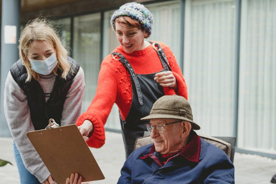 In Daylight two women stand outside looking at a clipboard held by an man  who is sat down. One woman points at the clipboard and smiles.