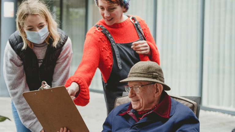 In Daylight two women stand outside looking at a clipboard held by an man  who is sat down. One woman points at the clipboard and smiles.