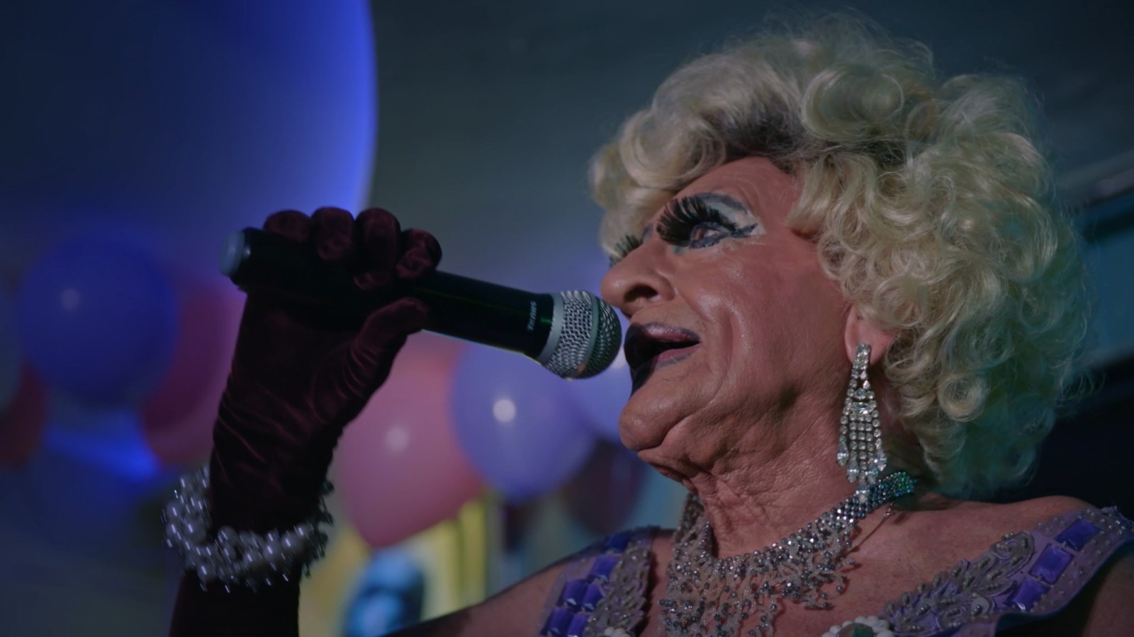 Elderly drag queen singing into microphone looking emotional