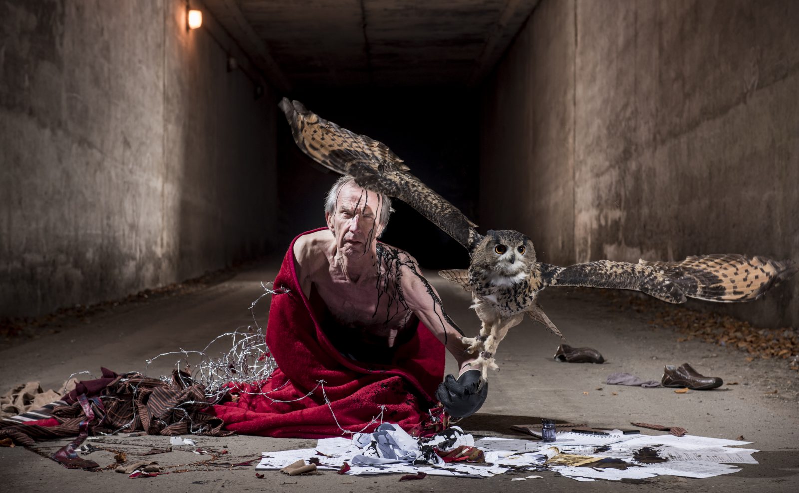 A topless man wrapped in a red blanket and barbed wire is kneeling in a concrete tunnel.  He is surrounded by clothes and papers. Oil drips on his head and down his shoulder. A large owl is taking flight from his outstretched arm.
