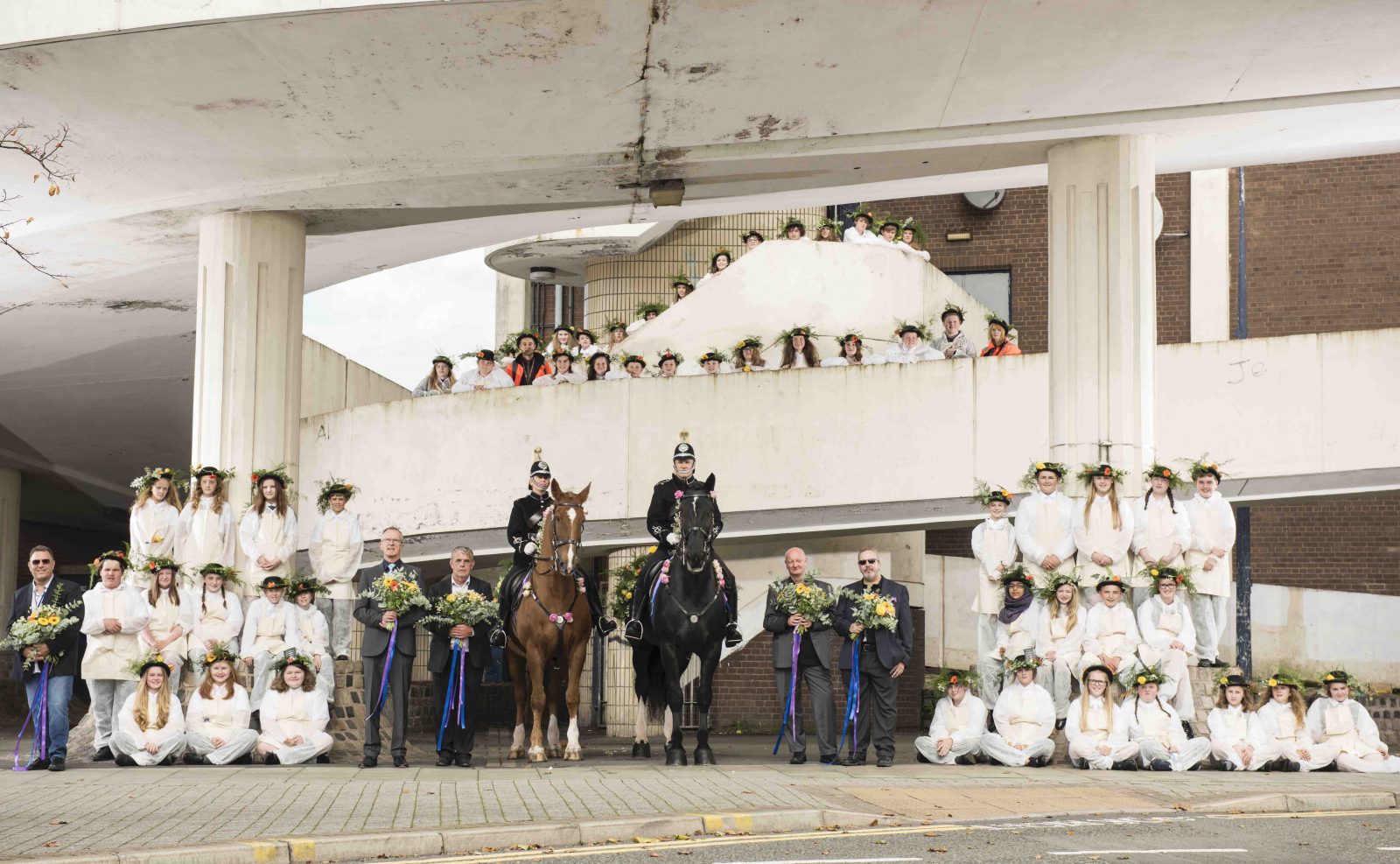 A very large group of people, around 60, are posed for a photograph on different levels of a walkway. Many of the people are young people, wearing white overalls, aprons and hates covered with flowers and greenery. 5 men wear suit jackets and trousers and hold large bunches of flowers. In the centre, on the ground level, are two police officers on horses wearing roses and flowers on their harnesses.
