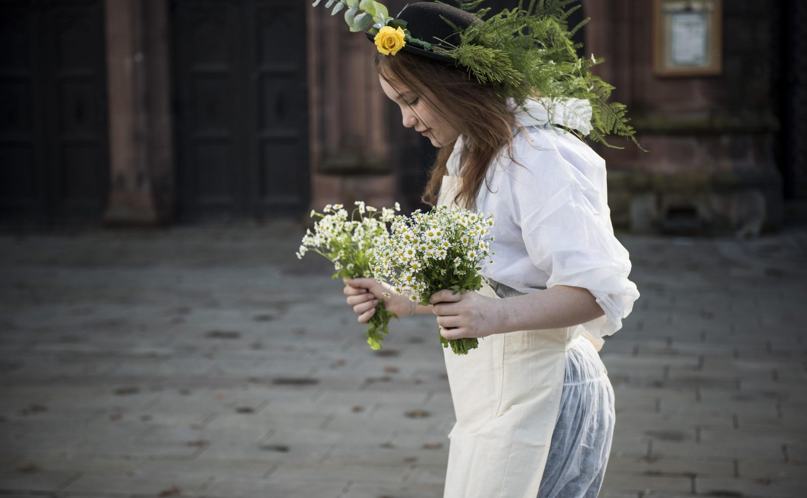 A young person walks in front of a sandstone building wearing white overalls and an apron. They are carrying two big bunches of white flowers and they wear a hat covered in flowers and green ferns.
