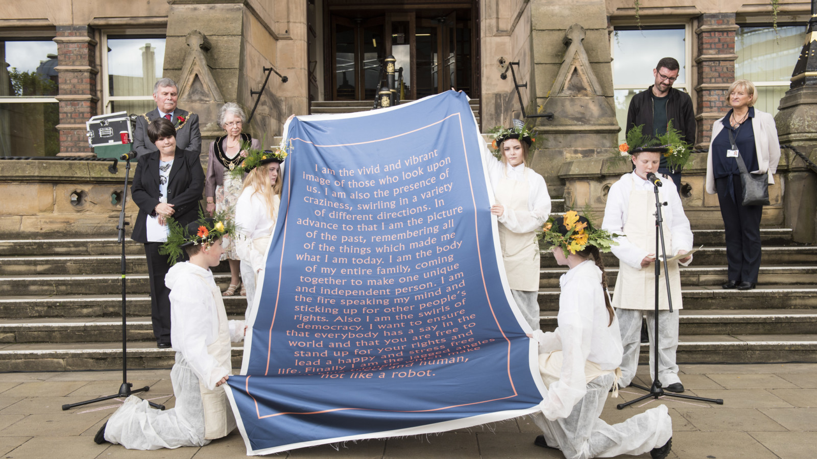 Four young people in white overalls and hats covered in flowers and greenery hold a large banner with writing on it. Another young person in the same outfit stands to one side with a microphone and is reading from a piece of paper. A number of adults stand nearby and are looking at the young person with the microphone.