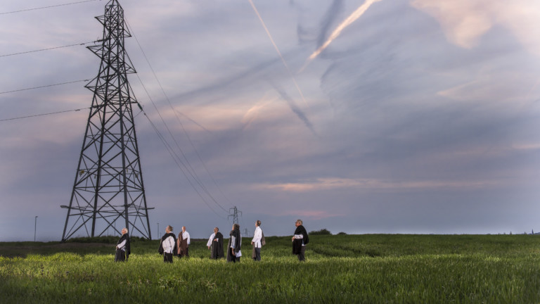 Seven men stand in a field of green grass, under a blue and grey dusky sky. There is a large electricity pylon in the field which towers over the men.
