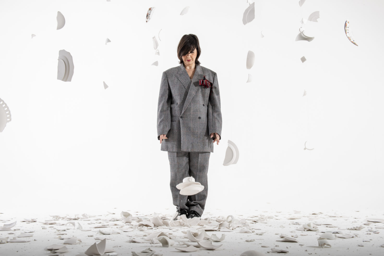 A woman stands in an all white space with smashed white crockery on the ground and falling around her. She wears an oversized grey suit and she looks down towards the ground at her feet.