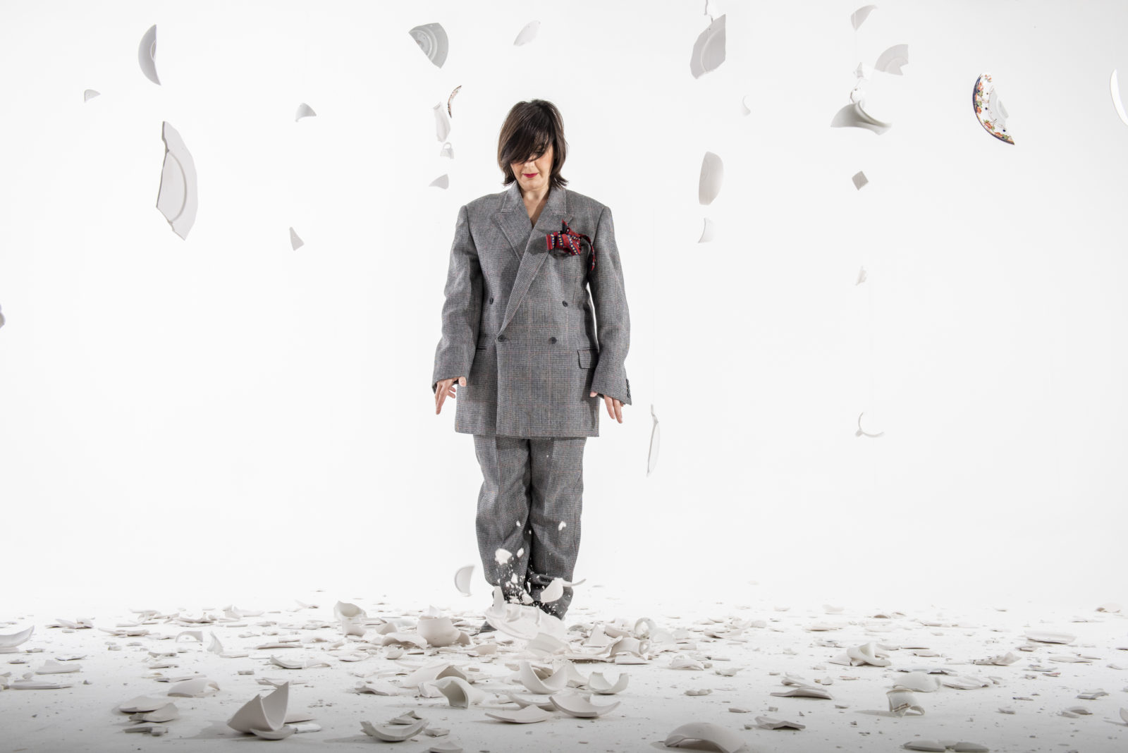 A woman dressed in a man's suit stands amongst broken crockery