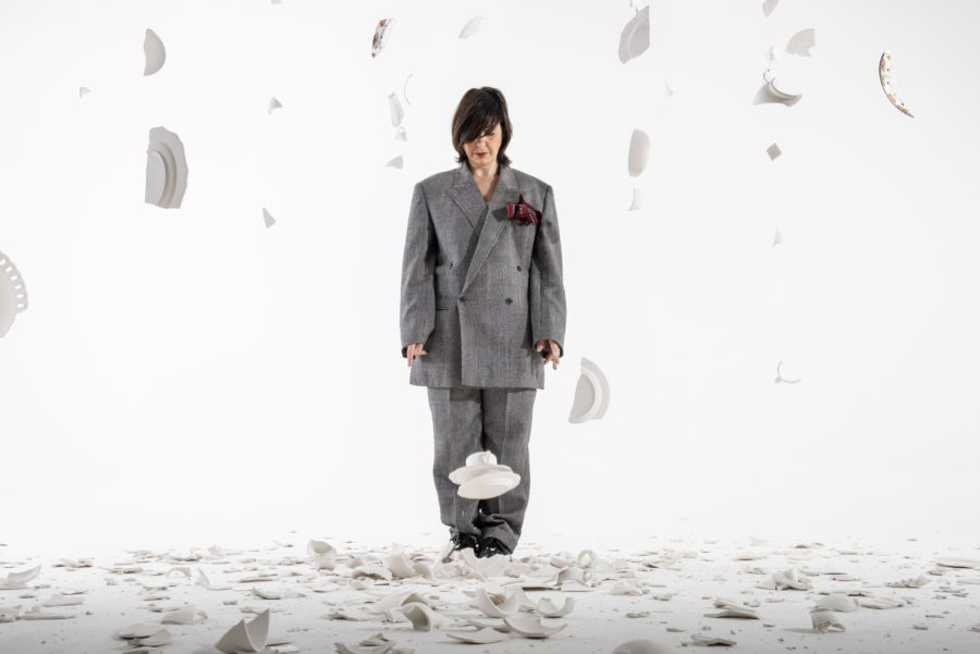 A woman stands in an all white space with smashed white crockery on the ground and falling around her. She wears an oversized grey suit and she looks down towards the ground at her feet.