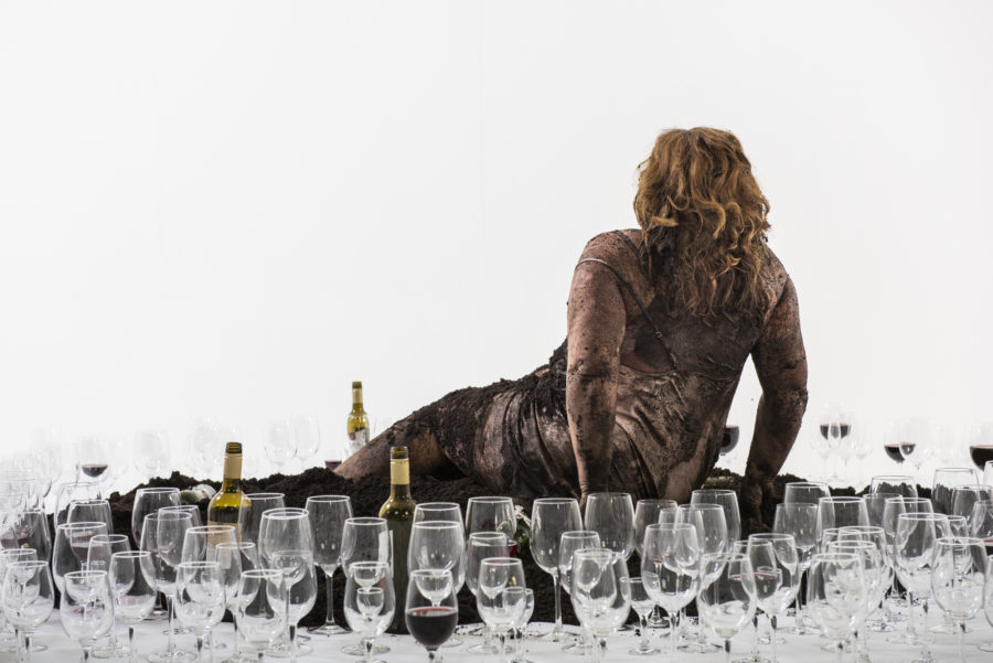 A fair skinned woman with long blonde hair sits on the floor in a pile of dirt, she wears a pink silk slip. She has her back to us, with her legs out-stretched, her body is smeared black with dirt. She is surrounded by bottles of wine and wine glasses, many empty, some full.