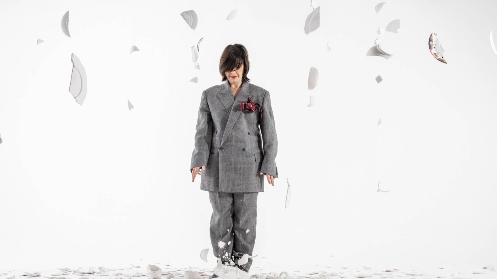 A woman dressed in a man's suit stands amongst broken crockery