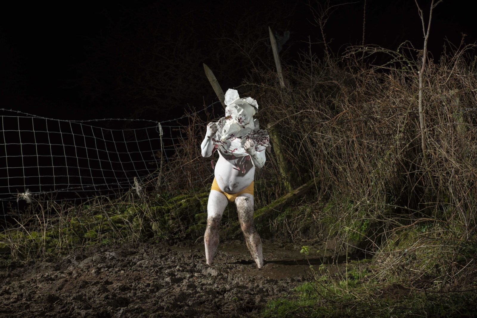 A man is outside in the dark in a muddy field, there is a barbed wire fence and bushes and branches behind him. He is covered in white powder and wearing yellow boxers, and on his head he is a bear shaped mask made of paper, covering his head and shoulders.