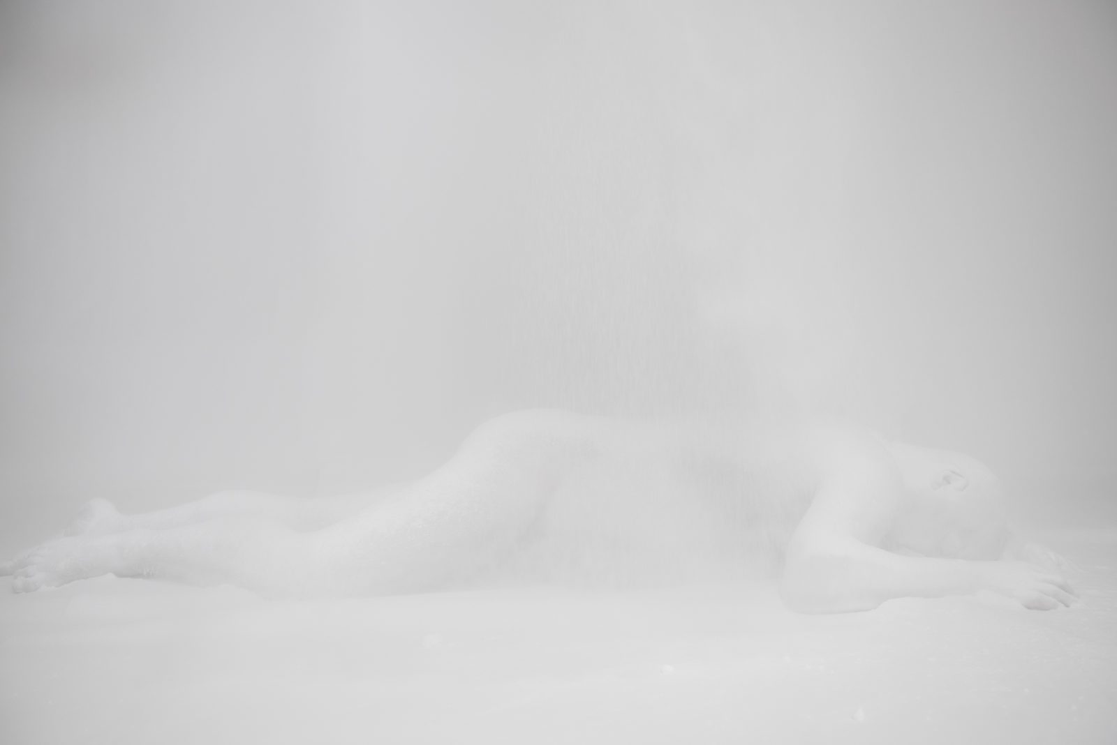 A man is lying down on his side in an all white room. He is covered in white powder, and the room is cloudy with the white poweder so you can only just see him.