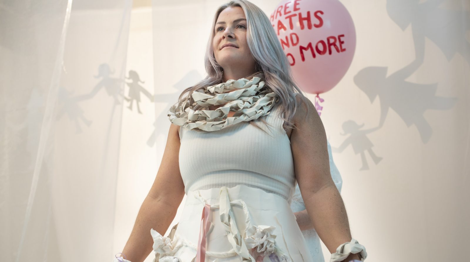 A woman with platinum hair stands upright with her arms by her side in a confident stance. She is smiling slightly and is wearing all white with some paper chains around her neck. Behind her are a string of paper dolls.