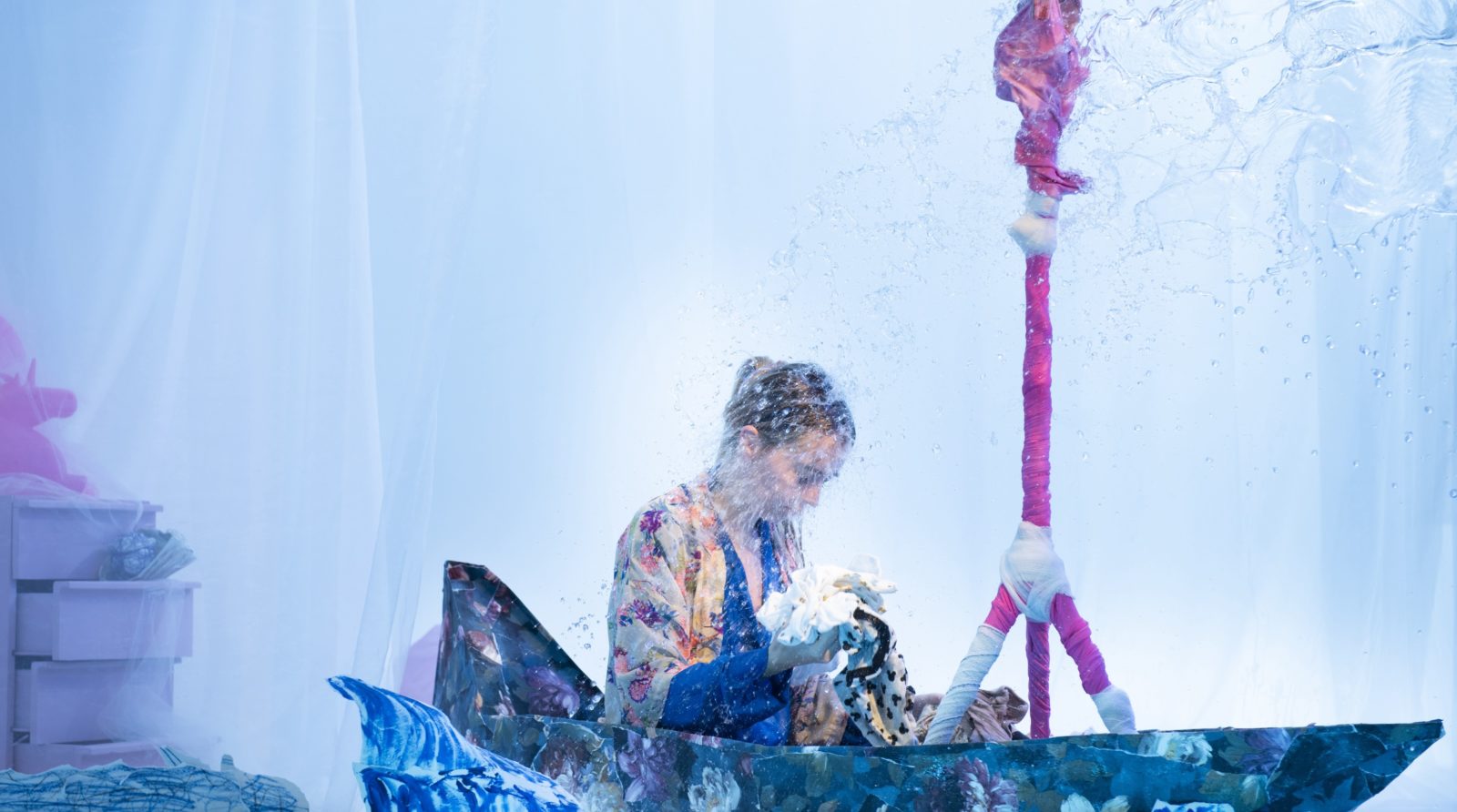 A woman in a boat that is made of floral printed paper, looks down at some children's clothes in her hands. Her expression is solemn.