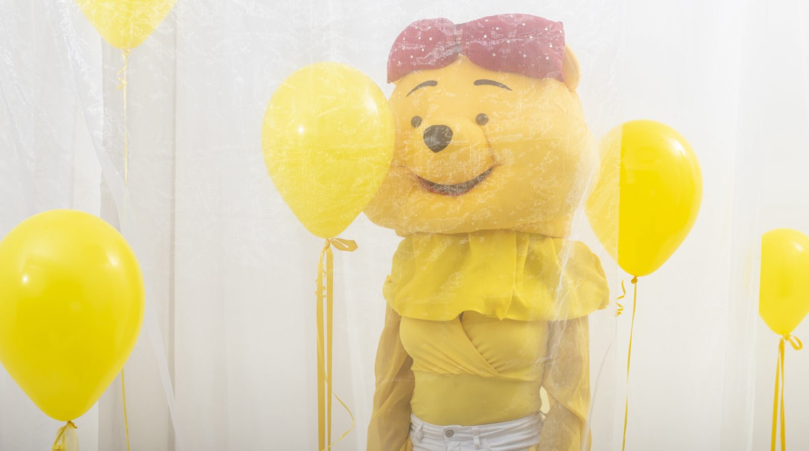 A person stands behind a piece of white netting, wearing a yellow top and a yellow bear mascot hat on their head. They are surrounded by yellow balloons.