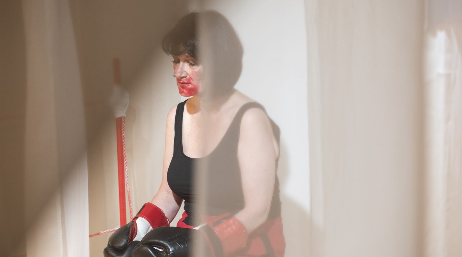 A woman with short brown hair is photographed wearing boxing gloves and has red lipstick smudged all around her mouth. She looks down and her expression is sad.