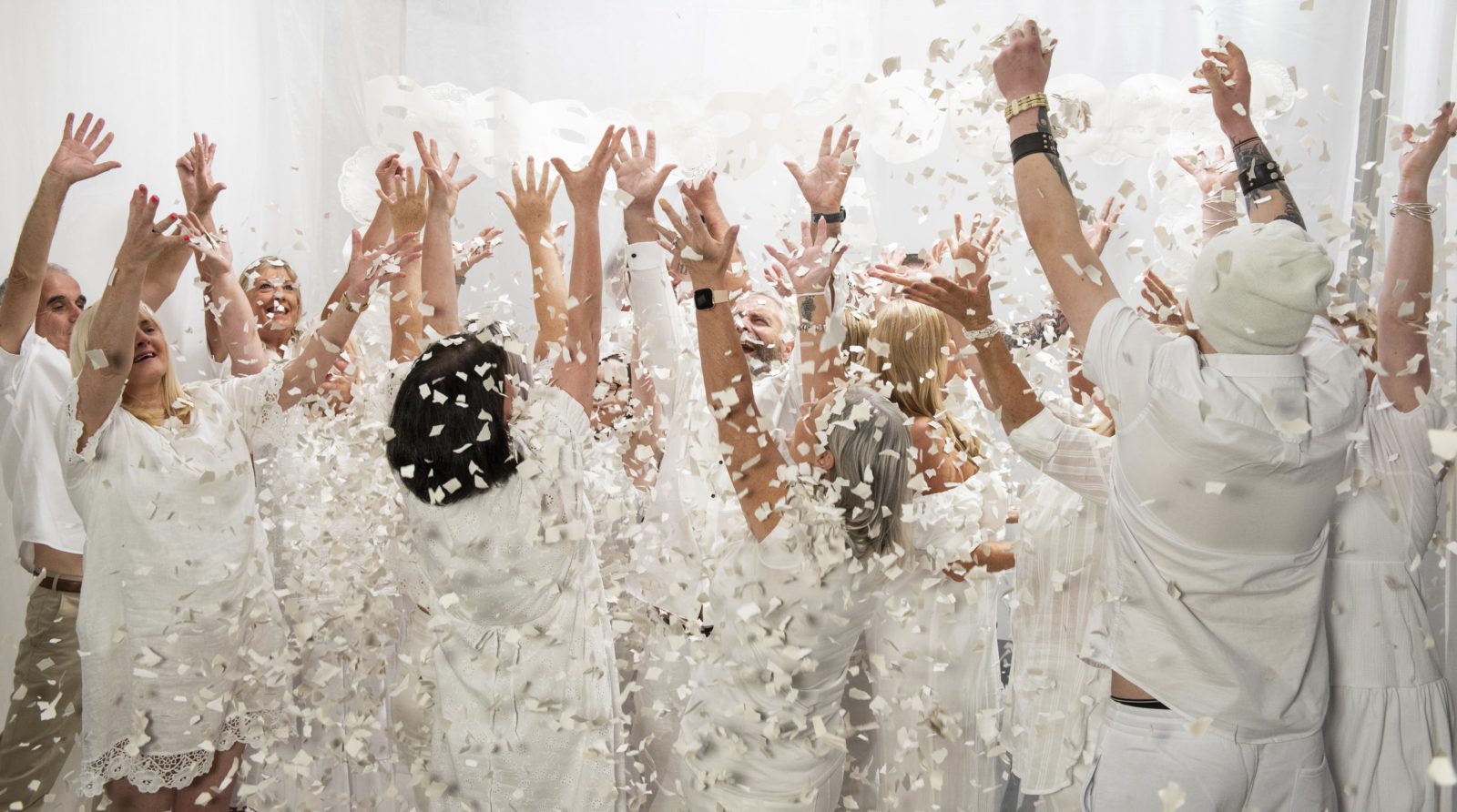 A group of people who are dressed all in white, are in a white room and throw white confetti in the air.