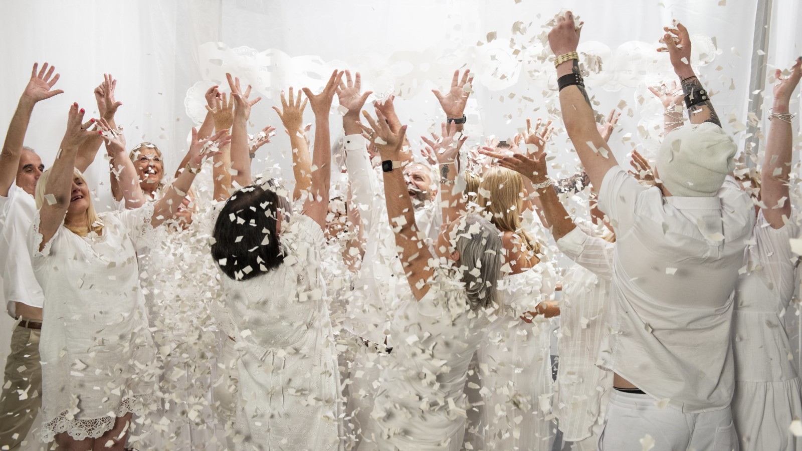 A group of people who are dressed all in white, are in a white room and throw white confetti in the air.