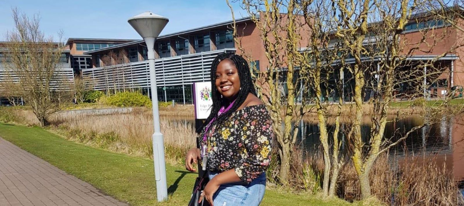 A black woman is pictured outside on a sunny day. Behind her a large ornamental pond surrounds a part of a large three story building. She is smiling, her dark top is patterned with flowers, and she wears blue jeans.