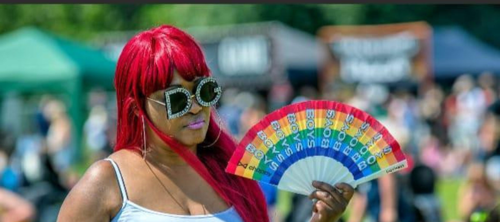A black woman with long, red-orange hair, wears a white vest top and sunglasses. She is holding a fan coloured with the rainbow like the LGBT+ flag.