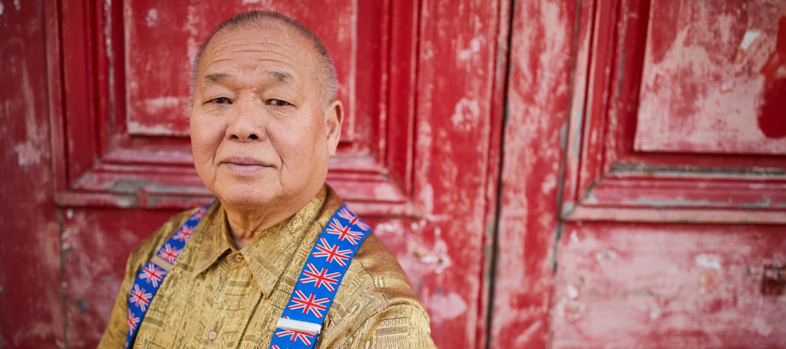 Sam, a 68 year-old Malaysian man, stands in front of a large red door. Sam wears a gold coloured shirt and union-jack braces.