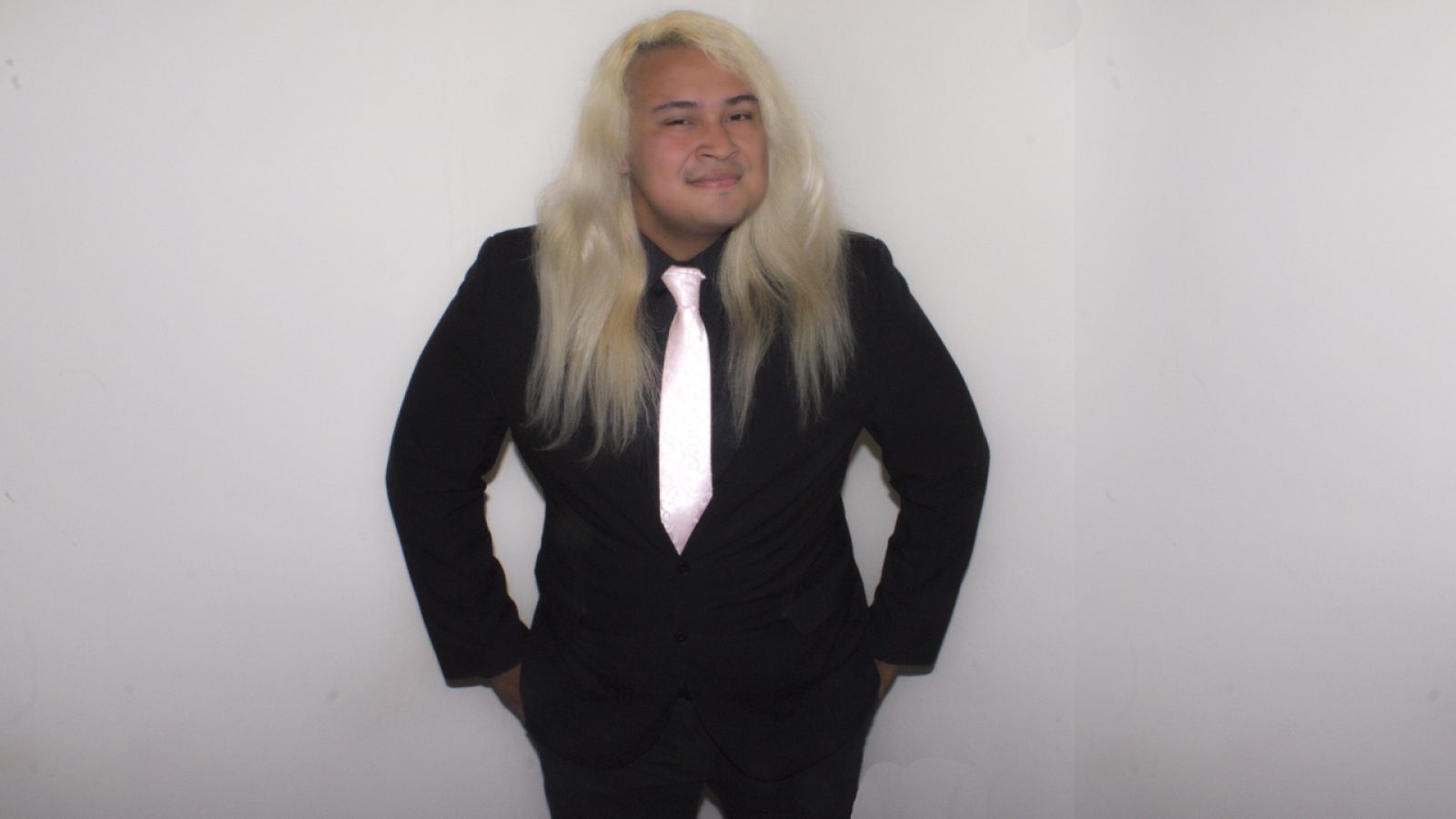 A young, light skinned man with long, blonde, wavy hair and brown eyes, wearing a black suit and shirt with a pink silk tie stands against a light grey background. He faces us with hands in trouser pockets, he has a content smile