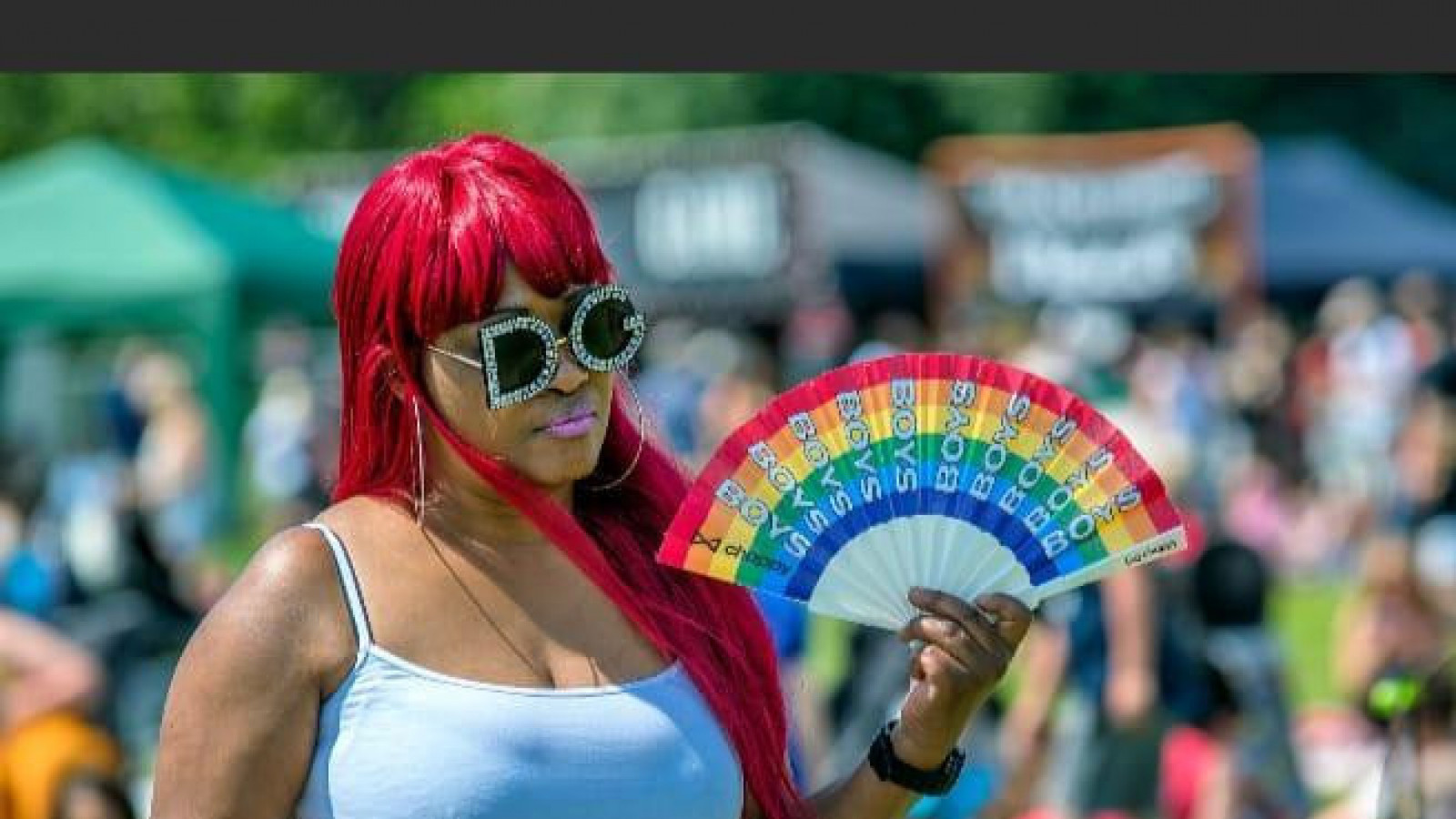 A black woman with long, red-orange hair, wears a white vest top and sunglasses. She is holding a fan coloured with the rainbow like the LGBT+ flag.