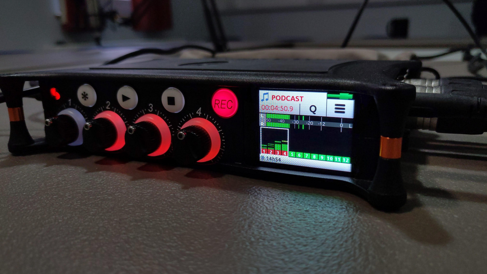 Recording equipment placed on a table. Its digital display is a small white screen containing information in black, green and red. The machine's buttons are illuminated red.
