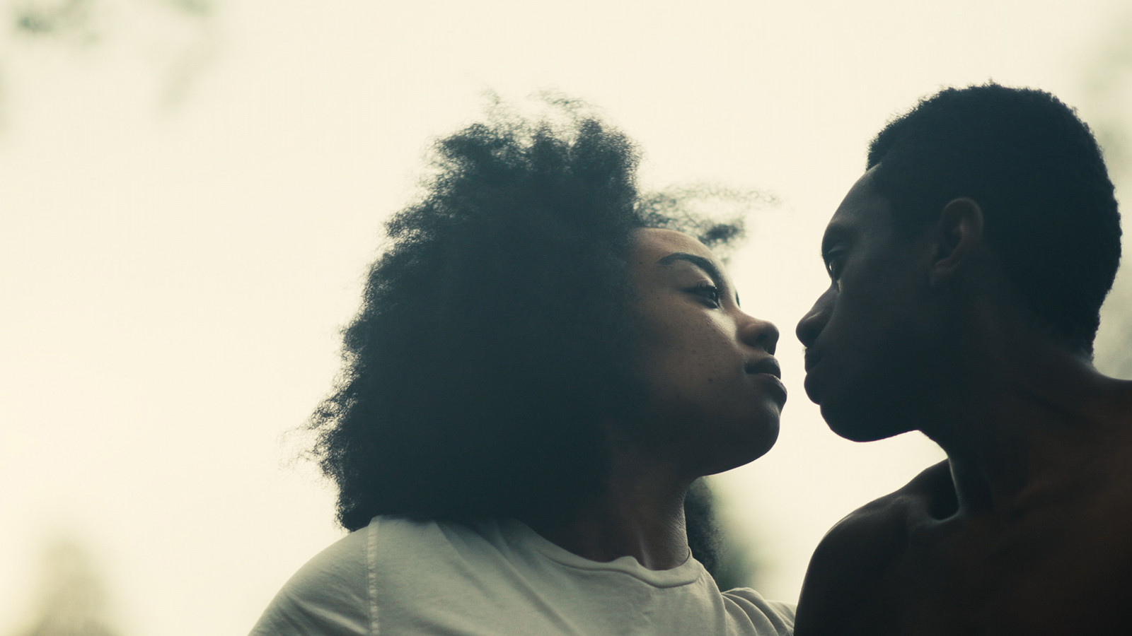 A dark skinned woman and man gaze into each other's eyes, their lips are nearly touching.