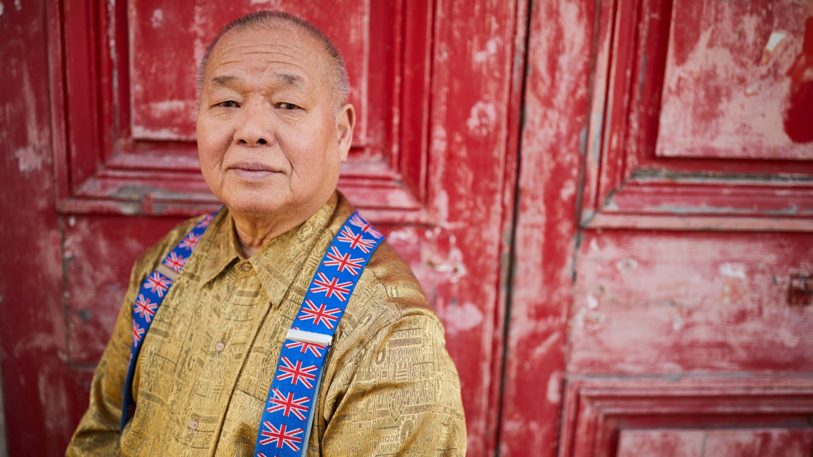 Sam, a 68 year-old Malaysian man, stands in front of a large red door. Sam wears a gold coloured shirt and union-jack braces.